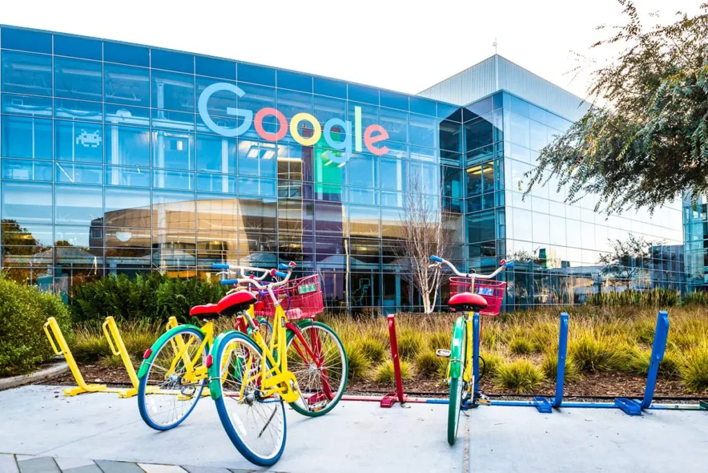 Fargerike sykler parkert utenfor en Google-partnerbygning med en stor glassfasade og Google-logoen.