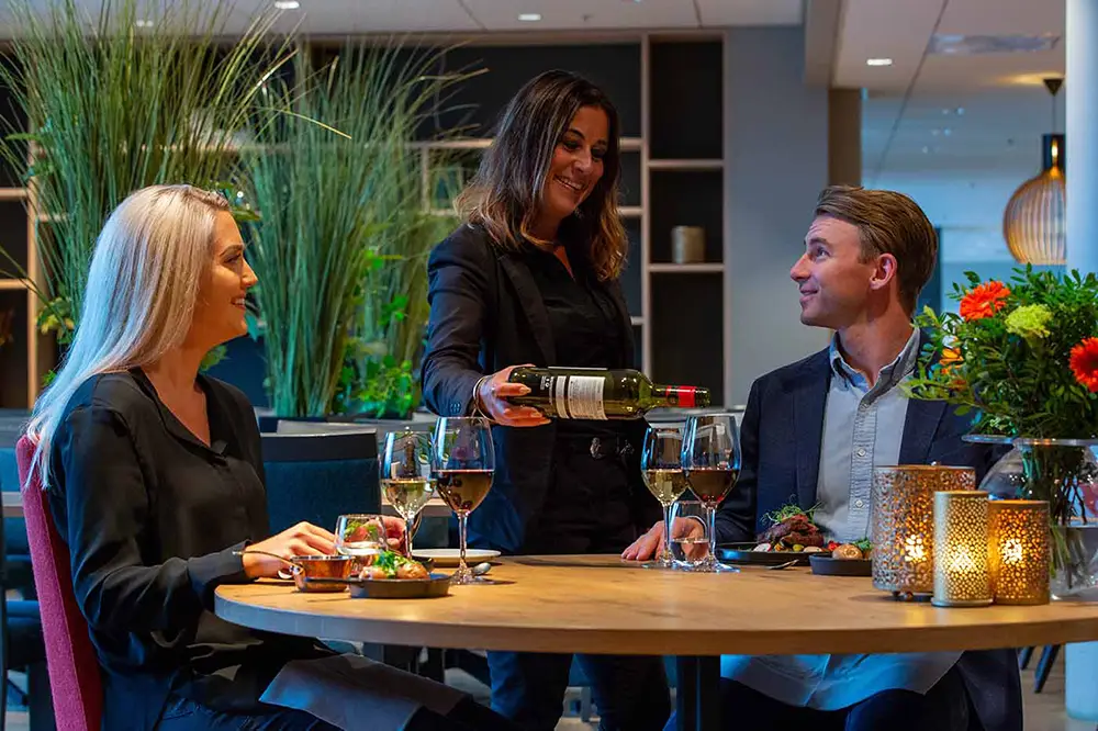 En servitør skjenker vin til en smilende mann og kvinne som sitter ved et Nye Elgstua restaurantbord med måltider og levende lys.