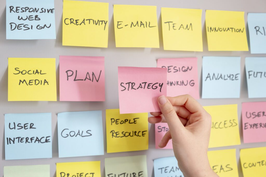 a person holding a piece of paper in front of a wall of sticky notes with marketing strategies for 2023.