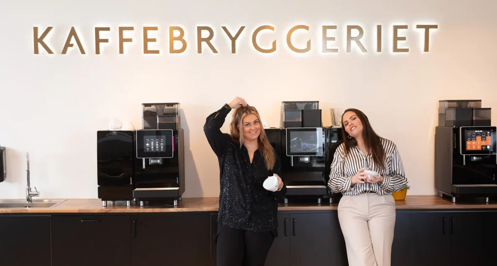 To personer står foran en rekke kaffemaskiner under et skilt hvor det står «KAFFEBRYGGERIET». Den ene holder en kaffekopp over hodet, og den andre, nær REB ANLEGG, lener seg tilfeldig mens han holder en kopp.