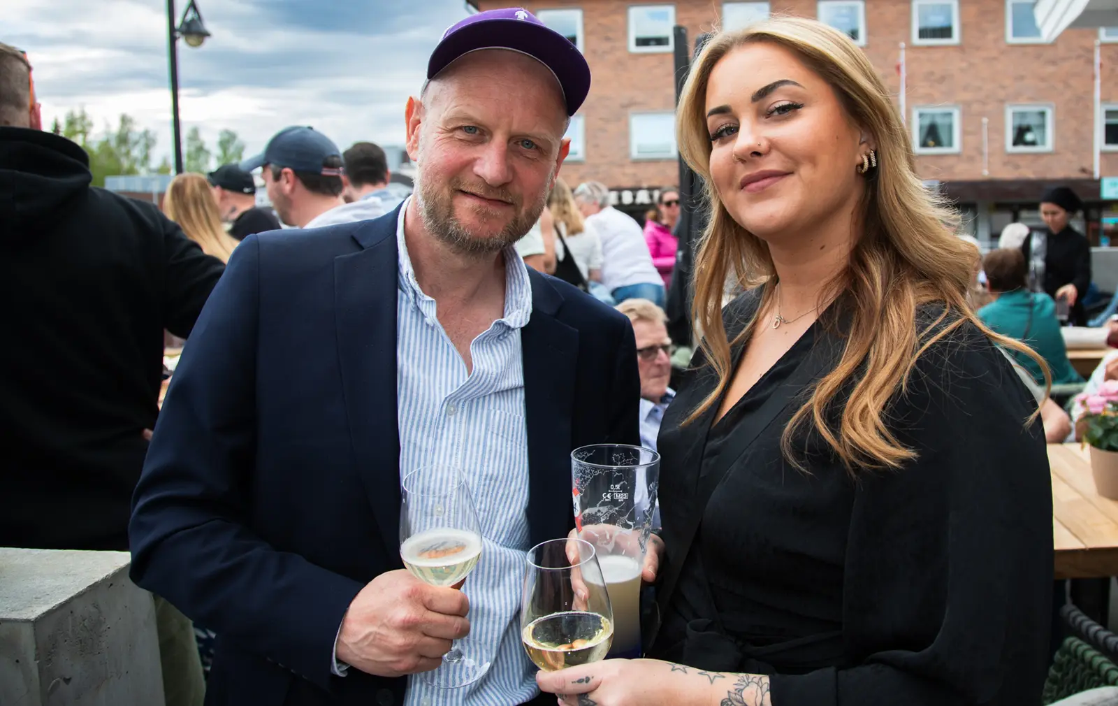 Victoria Moen Toth fanger et gledelig øyeblikk mens en mann og kvinne nyter drinker på en utendørs sammenkomst, mens de holder et glass og smiler til kameraet.