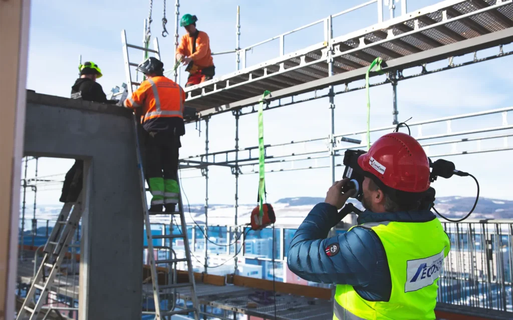 Bygningsarbeidere i verneutstyr jobber iherdig på et stillas, mens en arbeider dokumenterer prosessen ved hjelp av et kamera for en rekrutteringsfilm.