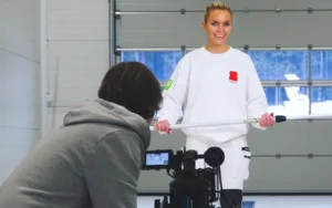 En kvinne i en hvit genser holder en lang hvit gjenstand mens hun står innendørs. En kameraoperatør i en grå hettegenser filmer henne og fanger scener for rekrutteringsfilmen.