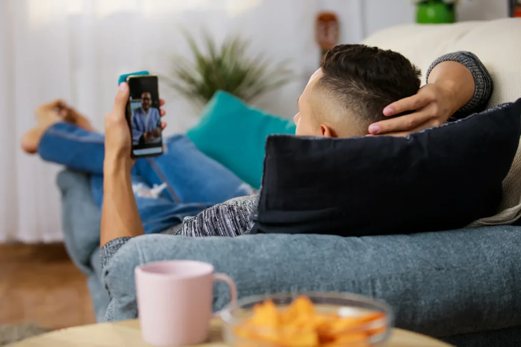 En person lener seg tilbake på en sofa, smarttelefon i hånden, engasjert i en videochat om salgsemner. Et krus og en bolle med snacks står på bordet i forgrunnen, og setter en avslappet scene for produktive samtaler.