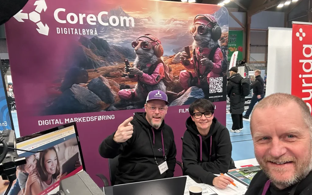 Tre personer på CoreCom-standen, med banner med tegneserieskapninger og teksten "Digitalbyrå" og "Digital Markedsføring." De smiler og engasjerer seg med kameraet, legemliggjør ånden i å lage engasjerende videoer for bedrifter.