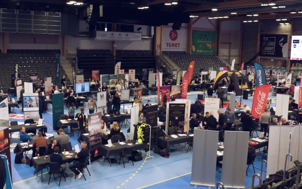 Vid utsikt over en jobbmesse i en stor gymsal, som minner om et Elverum Håndball nettverkstreff, med utallige stander, bannere fra ulike bedrifter og deltakere i samspill ved bord.