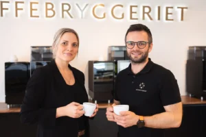 To personer som holder kaffekopper står foran en elegant disk som er utsmykket med toppmoderne kaffemaskiner. Bakteppet viser stolt "KAFFEBRYGGERIET" på veggen, og fanger essensen av skandinavisk bryggehåndverk.