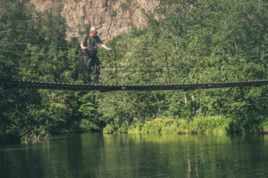 En person krysser forsiktig en hengebro over en svingete elv i en frodig skog, med de steinete klippene stående som standhaftige vaktposter. Med hvert trinn navigerer de både i naturens skjønnhet og de potensielle salgsleddene som venter utenfor horisonten.