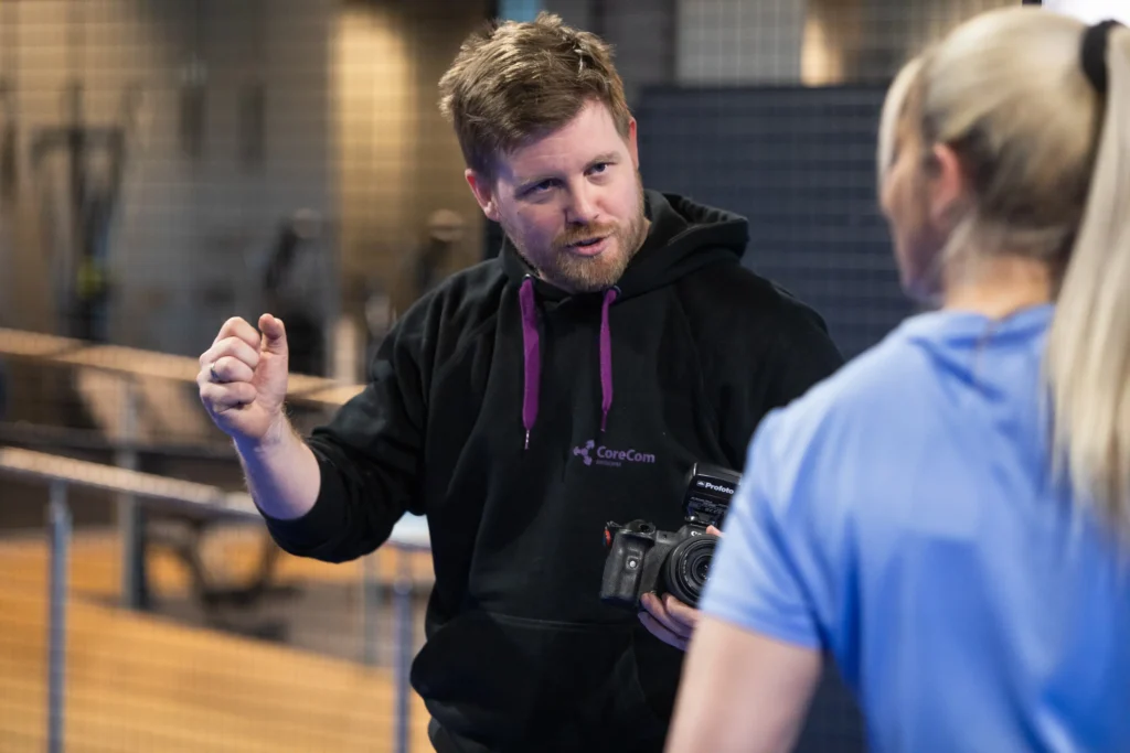 Anders Viken, som holder et kamera, snakker med en kvinne i et treningsstudio, og gestikulerer med hånden mens han vender mot henne.