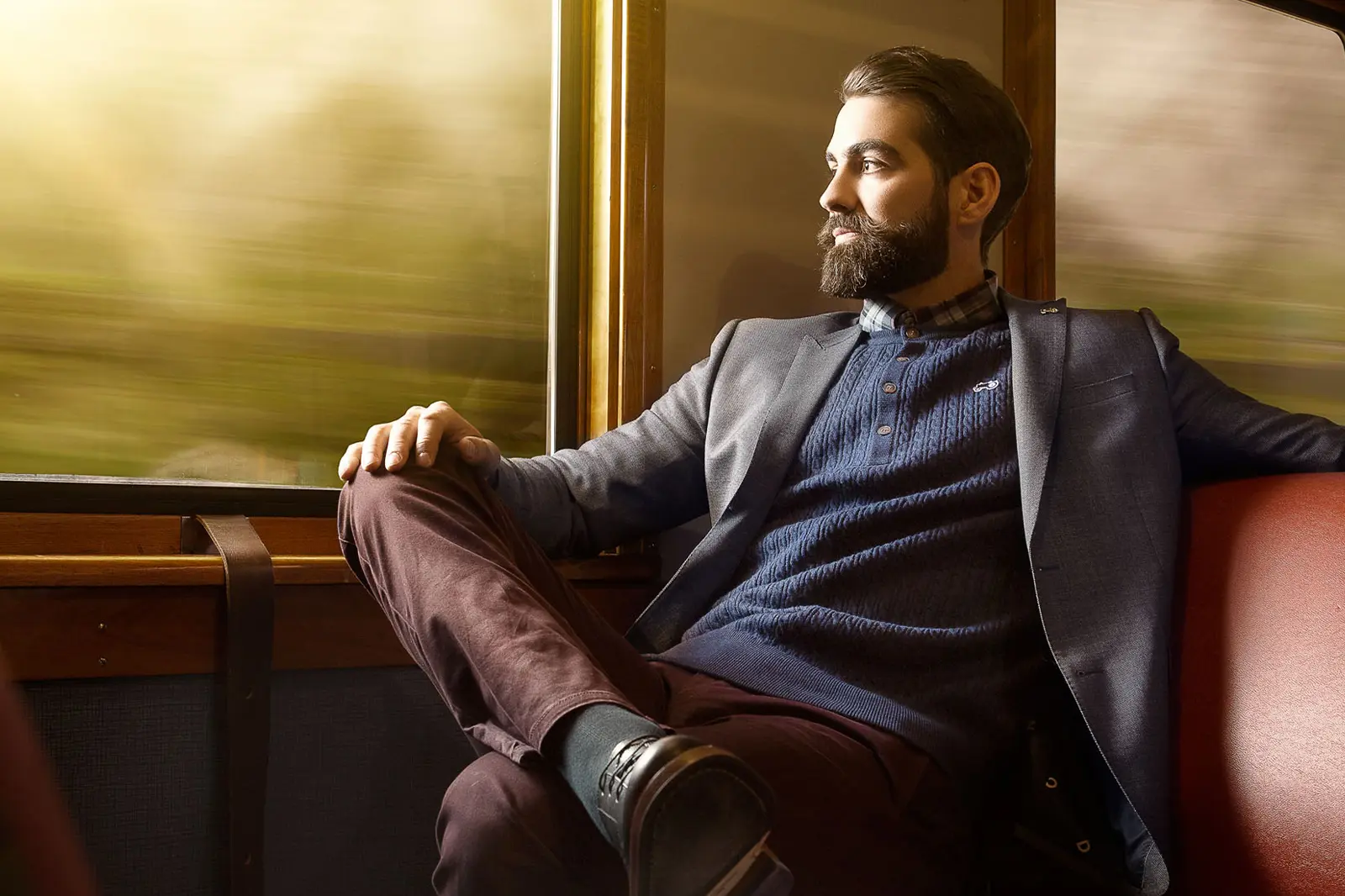 Anders Viken, en mann med skjegg, iført blazer og genser, sitter på et togsete og ser ut av vinduet på det uskarpe landskapet.