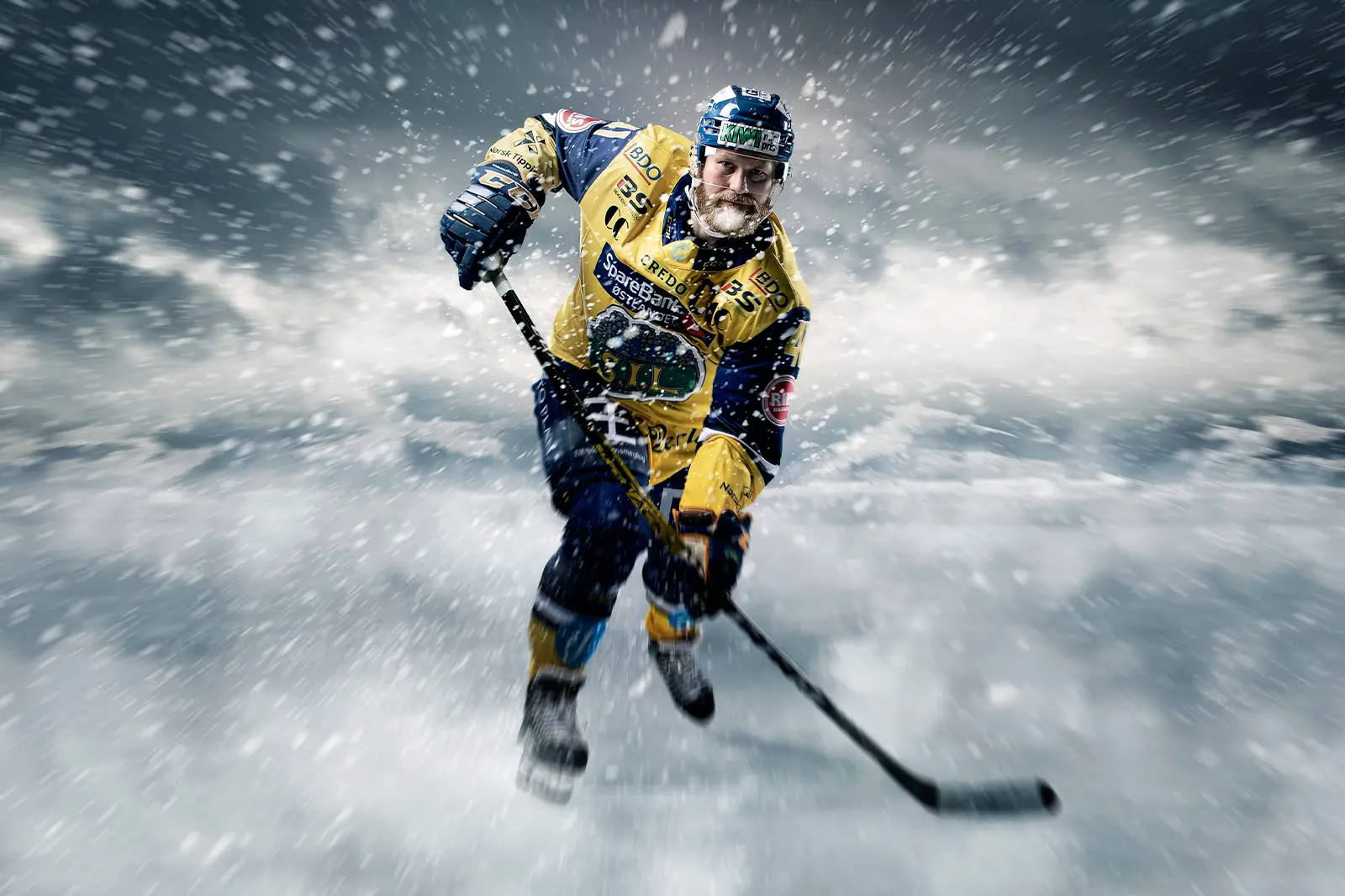 Ishockeyspilleren Anders Viken i gul og blå uniform skøyter over skøytebanen med en hockeykølle i hånden mens snø og is virvler i luften.
