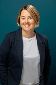 En kvinne med kort lysebrunt hår, iført en marineblå blazer over en hvit topp, står smilende foran en blågrønn bakgrunn.