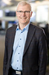 En eldre mann med briller, kort grått hår og en rutete blazer over en lyseblå skjorte står innendørs og smiler til kameraet.