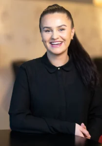 En kvinne med langt mørkt hår som er satt tilbake, iført en svart skjorte med knapper, smiler mens hun sitter ved et bord innendørs – perfekt for et moderne bedriftsfoto.