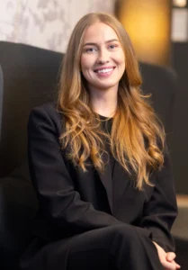 En kvinne med langt blondt hår, iført en svart dress, sitter på en mørk stol og smiler til kameraet innendørs – perfekt for et profesjonelt bedriftsfoto.