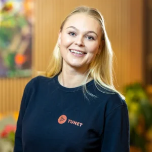 En kvinne med langt blondt hår smiler til kameraet, iført en mørk genser med en «TUNET»-logo. Bakgrunnen ser ut til å være et innendørsrom med planter og varm belysning.