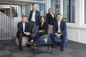 Fem profesjonelt kledde personer poserer for et gruppebilde i en moderne kontorlounge med store vinduer og et lite salongbord.