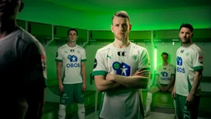 Fire mannlige fotballspillere i hvite og grønne uniformer poserer i en garderobe med grønn belysning, tre stående og én sittende på en benk.