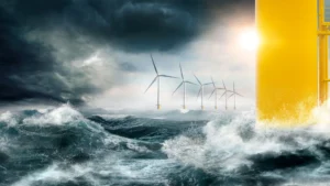 Offshore vindturbiner står i røffe havbølger under en stormfull himmel, med sollys som titter gjennom skyene og en gul struktur i forgrunnen.