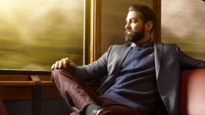 En mann med skjegg i blazer og genser sitter på et tog og ser ut av vinduet med uskarpt landskap i bakgrunnen.