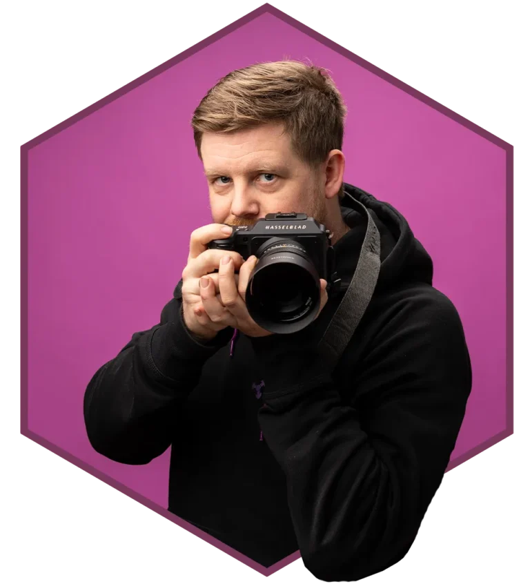 En person i svart hettegenser holder et Hasselblad-kamera opp mot ansiktet, stående mot en heldekkende lilla bakgrunn i en sekskantet ramme – perfekt for Corecom Digitalbyrås kreative visuelle merkevarebygging.