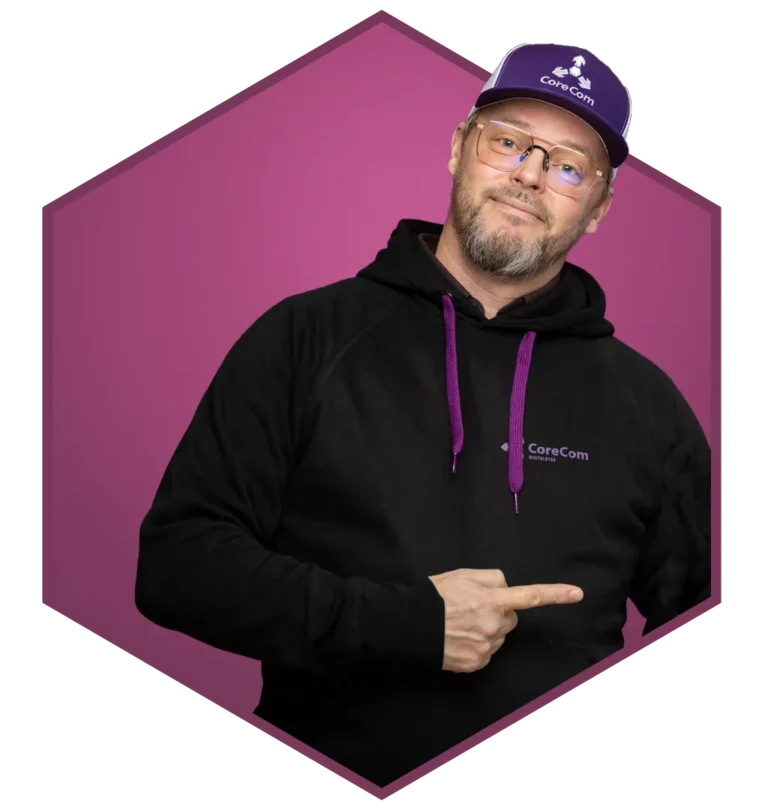 En person med briller, svart hettegenser med lilla aksenter og en lilla caps står mot en lilla sekskantet bakgrunn og peker på CoreCom-logoen på hettegenseren - perfekt til bruk i en rekrutteringsfilm.