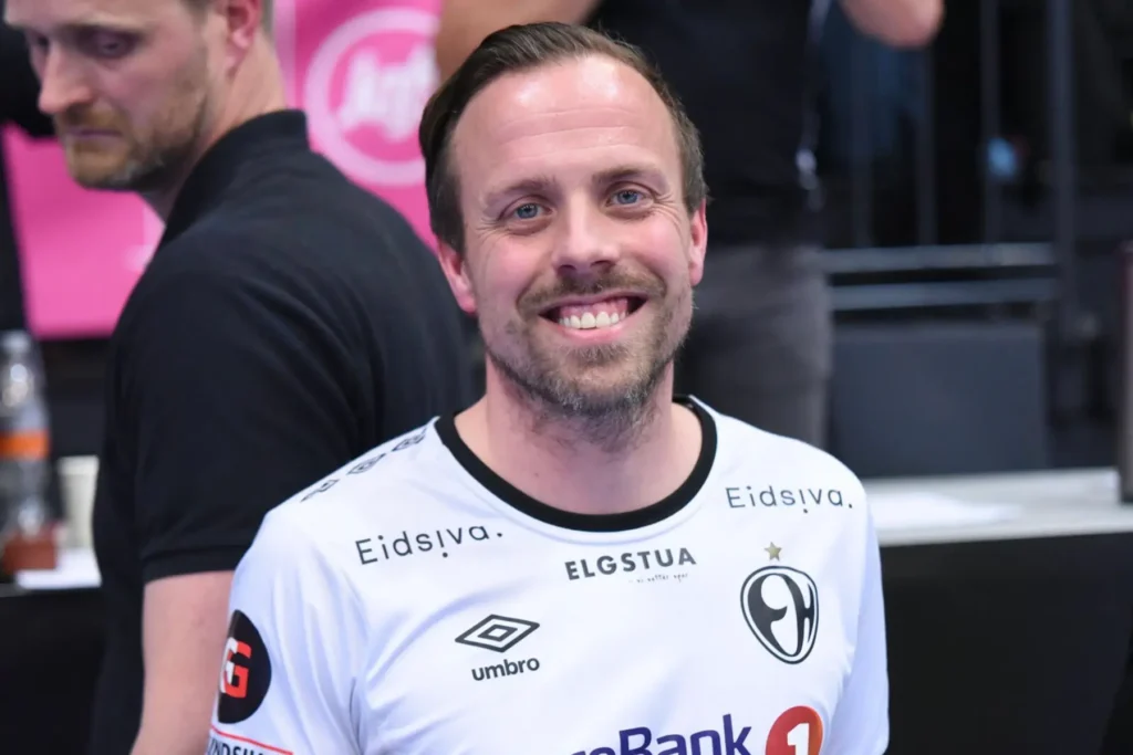 En mann i hvit sportstrøye med ulike sponsorlogoer smiler til kameraet innendørs, og viser sin entusiasme for å bygge salgsleads.