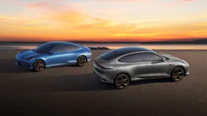 To moderne elbiler, en blå og en sølvfarget, står parkert på en åpen asfaltert flate i nærheten av et vann i solnedgangen - et ekte Bragd av innovativ design og bærekraft.