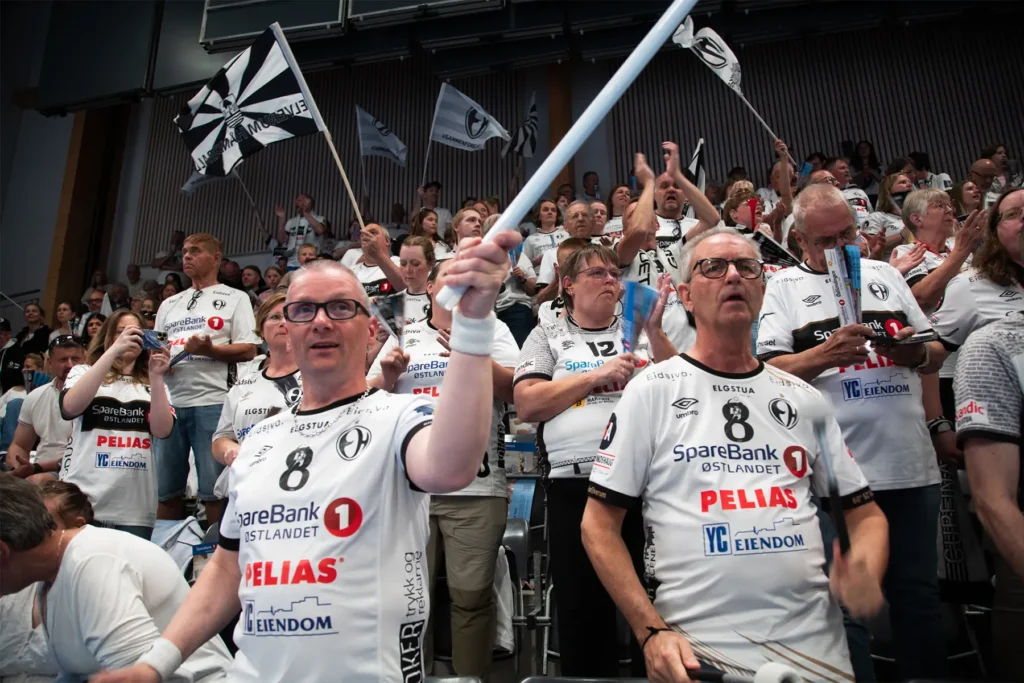 Fans i matchende hvite trøyer jubler og vifter med flagg i en fullsatt innendørsarena, og viser sin Salgsleads CoreCom-ånd mens de lidenskapelig støtter laget sitt under arrangementet.