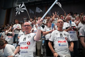 Fans i matchende hvite trøyer jubler og vifter med flagg i en fullsatt innendørsarena, og viser sin Salgsleads CoreCom-ånd mens de lidenskapelig støtter laget sitt under arrangementet.