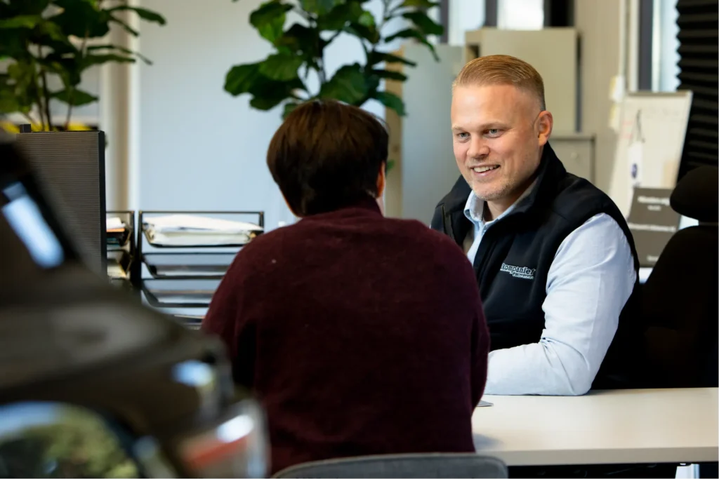 En mann og en kvinne sitter overfor hverandre ved et kontorpult, i samtale om Gillgruppen, med kontorplanter og papirarbeid synlig i bakgrunnen.