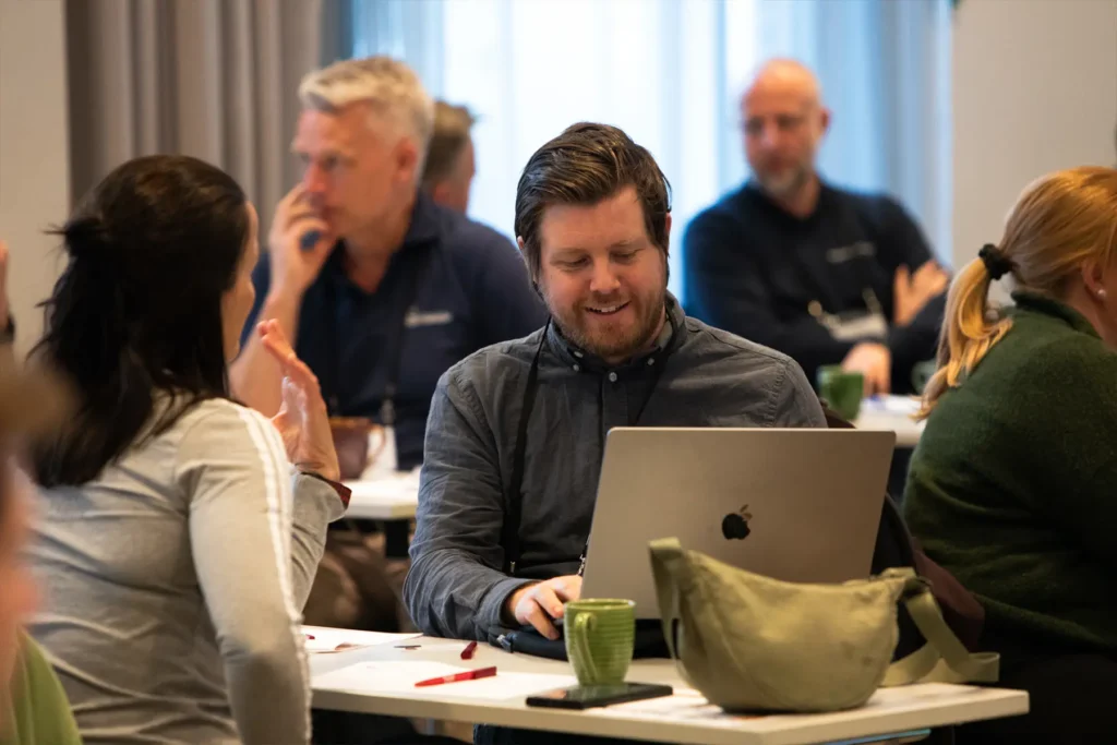En mann sitter ved et bord med en bærbar datamaskin og smiler, mens de andre rundt ham diskuterer nye strategier for å generere salgsleads i et CoreCom-konferanserom.