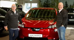 To personer står på hver sin side av en rød Subaru Solterra inne på Moss AutoCenter, begge iført svarte jakker og briller.