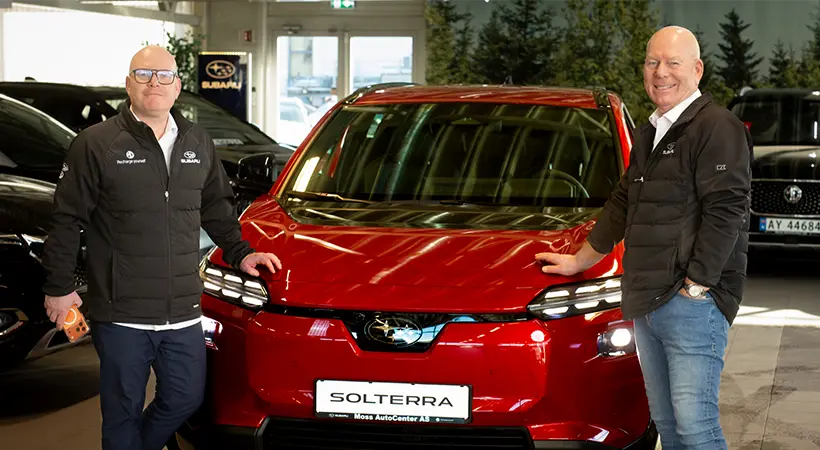 To personer står på hver sin side av en rød Subaru Solterra inne på Moss AutoCenter, begge iført svarte jakker og briller.